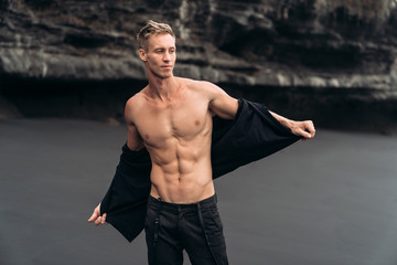 Sexy fitness male model in black pants and shirtless posing on black sandy beach.