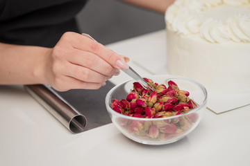 An attractive young female brunette confectioner decorates a white cake with small red flowers of food rose with tweezers.