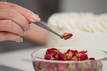 An attractive young female brunette confectioner decorates a white cake with small red flowers of food rose with tweezers.