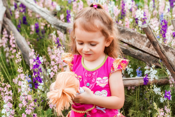 Cute baby girl in bright dress plays with doll outdoors in green field