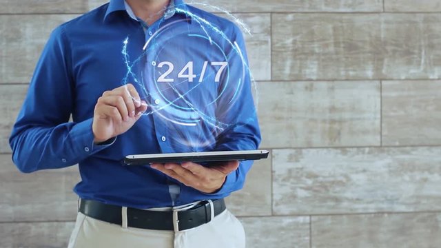 Man activates a conceptual hologram with text 24 7. The guy in the blue shirt and light trousers with a holographic screen gadget on the background of the wall