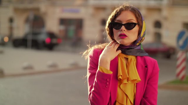 Outdoor portrait of fashionable woman with freckled skin, wearing black cat eye sunglasses, kerchief, colorful clothes, posing in street of european city