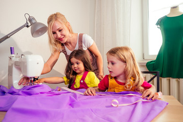 Smiling happy multitasking blonde mother teaches a little sisters to sew on a sewing machine by...