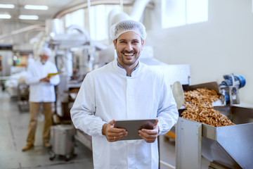 Young smiling manager in sterile uniform holding tablet and looking at camera while standing in food factory.