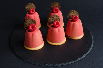 Set of chocolate mini mousse cakes covered with pink velvet spray, decorated with chocolate elements and fresh raspberry on black background