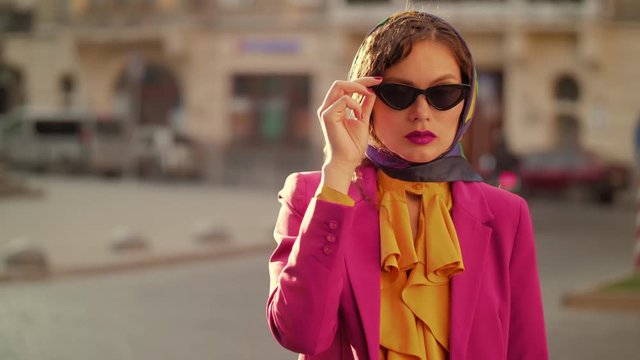 Fashionable woman wearing black cat eye sunglasses, kerchief, colorful clothes, looking at camera, posing in street of european city