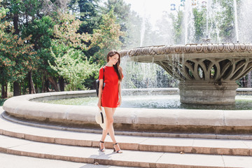 Young stylish beautiful brunette woman wearing red romper, black high heel sandals, black backpack and straw hat walking outdoors near fountain in the city. Trendy casual summer outfit. Street fashion