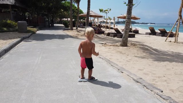 Cute two year old white boy trying to walk down the path in big female summer flip flops. beach in the background.