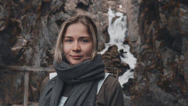 Close-up portrait: a beautiful young blonde girl in a scarf stands on the background of a waterfall, looking at the camera and smiling. Girl in a hike to a wild waterfall in the rocks. Slow motion, 4K