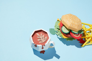 A cup of coffee with cheeseburger and french fries is made from paper craft on a blue background