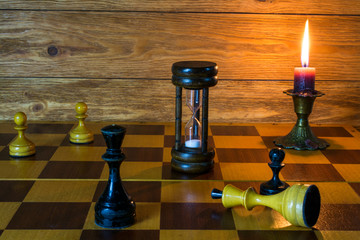 An hourglass, a burning candle and ches pieces  standing on a ches board.