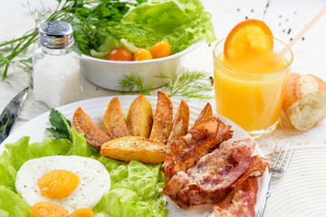 Homemade breakfast with potatoes with crispy bacon, tender egg and a glass of fresh juice on a white table background.