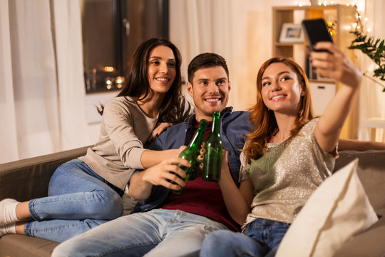 friendship and leisure concept - happy friends clinking drinks and taking selfie at home party in evening