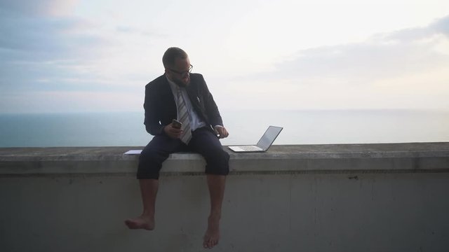 young businessman in a suit without shoes is sitting and working in a laptop on a concrete parapet on the background of a sea sunset