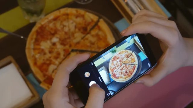 Hands of a woman or girl making a photo of a Hawaiian pizza on a restaurant table on a smartphone. Close-up