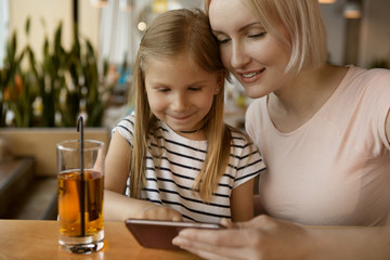 Front view of beautiful woman sitting with little child in cafe and watching video. Attractive mother and smiling girl drinking fresh juice and keeping phone. Concept of technology and relax.
