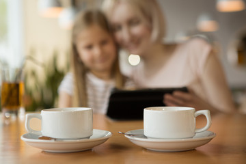 Two white cups of warm tea standing on table in cafe while mother and daughter sitting and watching video at background. Smiling woman and girl ordering cappuccino and coffee. Concept of lunch time.