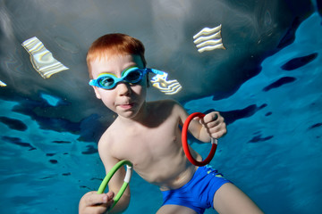 Cute sports boy swimming underwater in the pool with toys in his hands. Posing for the camera with...