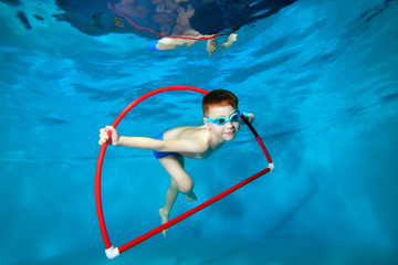 Smiling little boy, swimming in the pool, underwater at the bottom, swims through a red Hoop with toys in his hands and looking at the camera. Underwater photography. Horizontal orientation © alexbard
