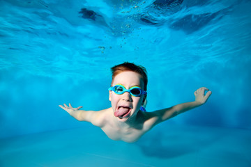 Happy smiling baby, boy, swims underwater in swimming pool in swimming glasses and shows tongue on camera on blue background. Portrait. Underwater photography. Horizontal orientation © alexbard