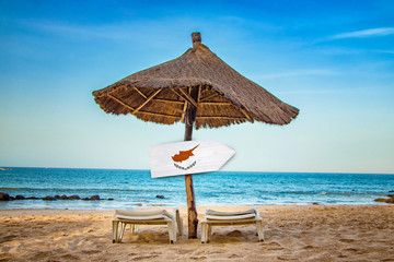 Cyprus flag on wooden arrow sign. There are two sun loungers and a sun umbrella on the beach. It is...