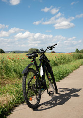 Fototapeta premium a bike, a bike with electric drive stands on a stone path next to the spring green field with red poppies. On far background, blue sky with white clouds.