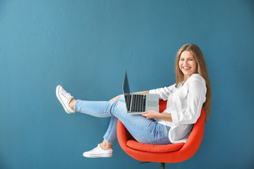 Beautiful young woman with laptop sitting on chair against color background - obrazy, fototapety, plakaty