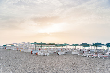 Lot of white deck chairs under an umbrella on a clean beach at sunset or sunrise.