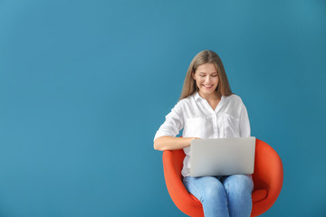 Beautiful young woman with laptop sitting on chair against color background