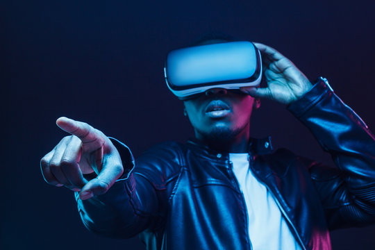 African guy pointing forward with finger while exploring virtual reality, pictured in darkness with neon light