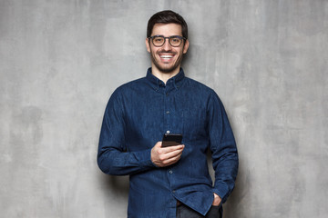 Positive young European man laughing at joke while holding smartphone, isolated on grey background