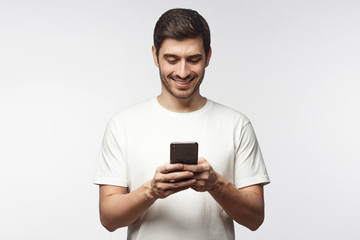 Studio portrait of young european caucasian man isolated on gray background wearing white t-shirt standing in front of camera, looking attentively with smile at screen of smartphone he is holding