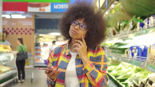 Beautiful african american woman with an afro hairstyle with smartphone in hand difficult to choose in the supermarket