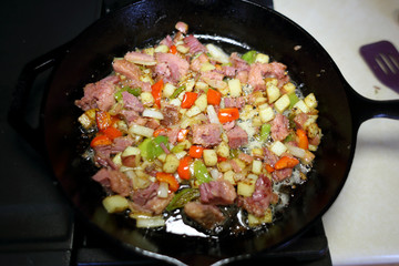 Corned beef hash frying in a cast iron skillet on a natural gas stove top in a home kitchen.