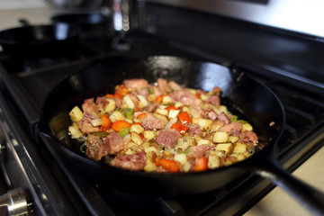 Corned beef hash frying in a cast iron skillet on a natural gas stove top in a home kitchen.