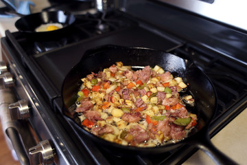 Corned beef hash frying in a cast iron skillet on a natural gas stove top in a home kitchen.