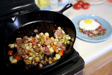 Corned beef hash frying in a cast iron skillet on a natural gas stove top in a home kitchen.