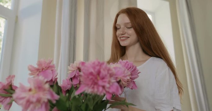 Attractive girl put the flowers of peonies in a vase and corrects the bouquet. Red-haired woman enjoying the fresh flowers and aroma of peonies. tracking shot. in slow motion. Shot on Canon 1DX mark2 