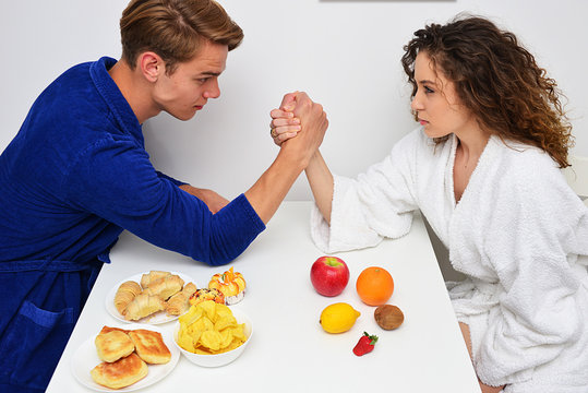 Young Couple Sitting At Table Arguing Over Healthy And Unhealthy Meal, Junk Food Over Fresh Fruit, Beautiful Girl And Boy Model