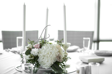 Wedding table setting is decorated with fresh flowers and white candles. Wedding floristry. Bouquet with roses, hydrangea and eustoma. On the background blur are burning garlands with light bulbs.
