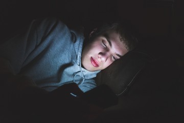 Teenager lying down on a couch in the dark. The light from the screen of his smartphone is illuminating his face.