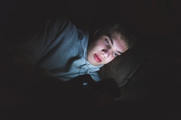 Teenager lying down on a couch in the dark. The light from the screen of his smartphone is illuminating his face.