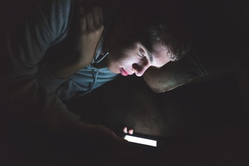 Teenager lying down on a couch in the dark. The light from the screen of his smartphone is...