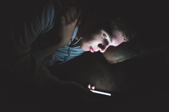 Teenager lying down on a couch in the dark. The light from the screen of his smartphone is illuminating his face.