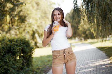 Beautiful young woman drink orange juice and walking in summer park.