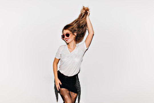 Graceful young woman in trendy pantyhose plays with long brown hair. Indoor photo of laughing ecstatic girl in cute sunglasses and stylish t-shirt relaxing on photoshoot.