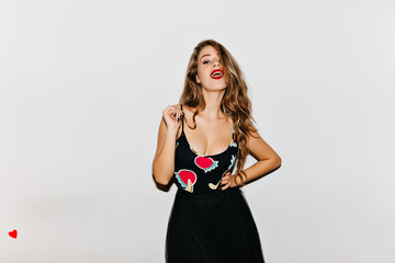 Fototapeta premium Stunning tanned woman in black dress standing in confident pose. Indoor portrait of wonderful european girl with bright makeup isolated on light background.