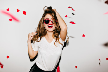 Indoor portrait of amazing caucasian female model in trendy t-shirt touching her long shiny hair. Laughing refined woman in sunglasses having fun with red confetti. - obrazy, fototapety, plakaty