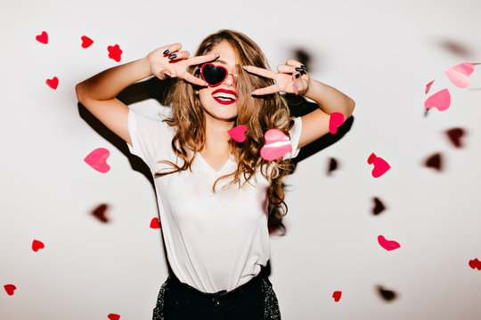 Indoor photo of stunning european lady in red sunglasses having fun at home party. Lovely girl in white t-shirt fooling around under heart confetti.