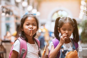 Two cute asian child girls with backpack eating pancake together after school in the school